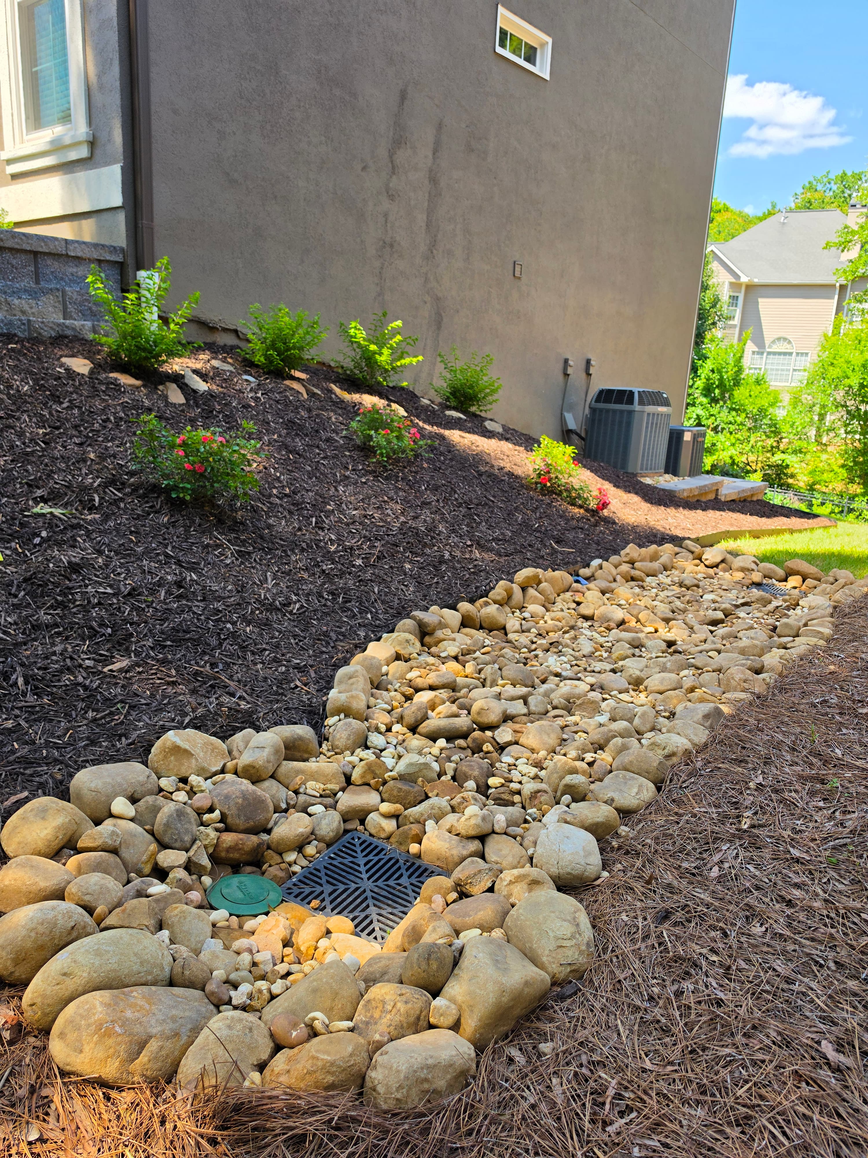 Landscaped river rock dry creek bed and drainage grate beside a mulched garden slope.
