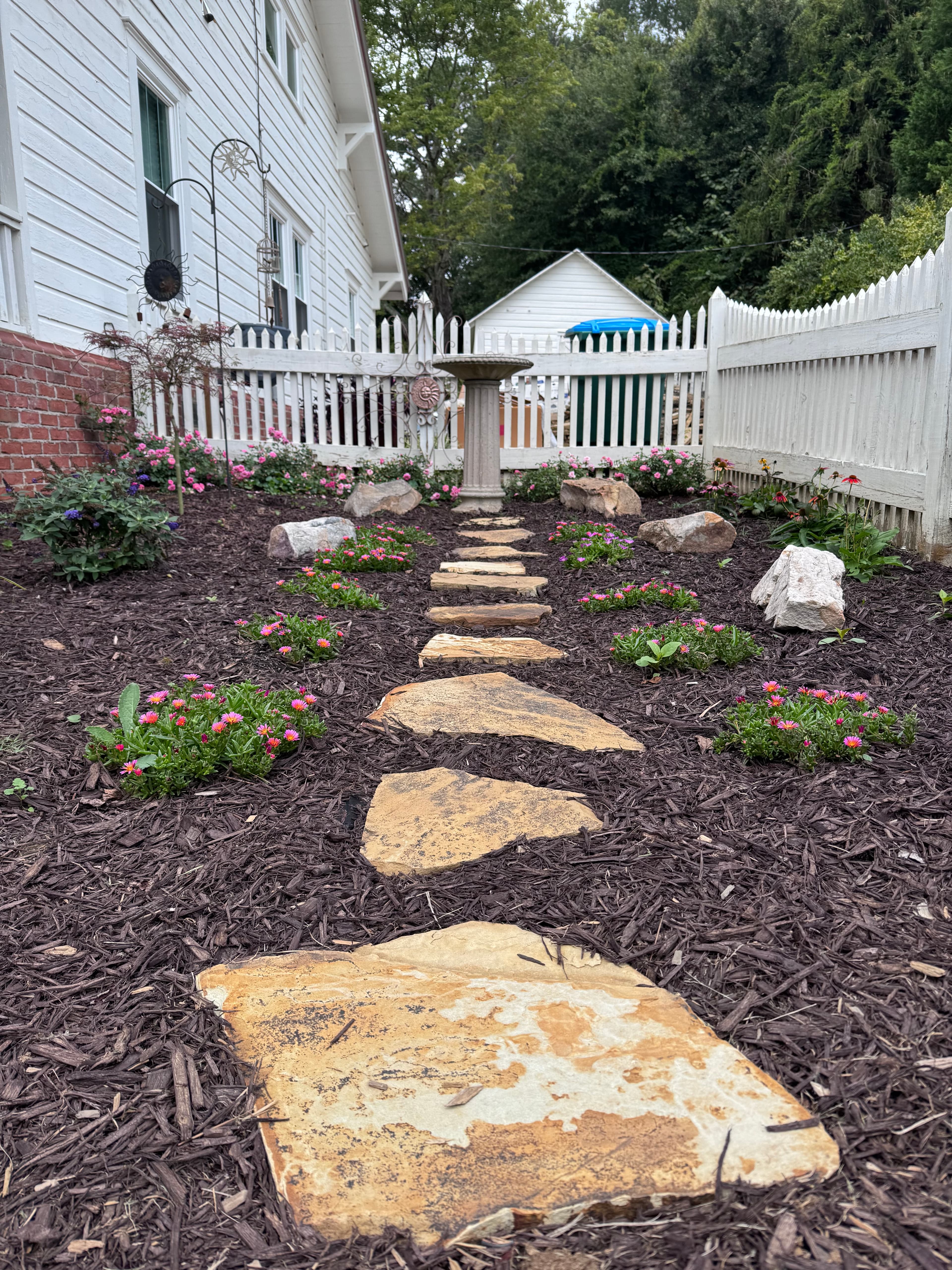 Tan stepping stone path through dark mulch and pink flowers along white picket fence.