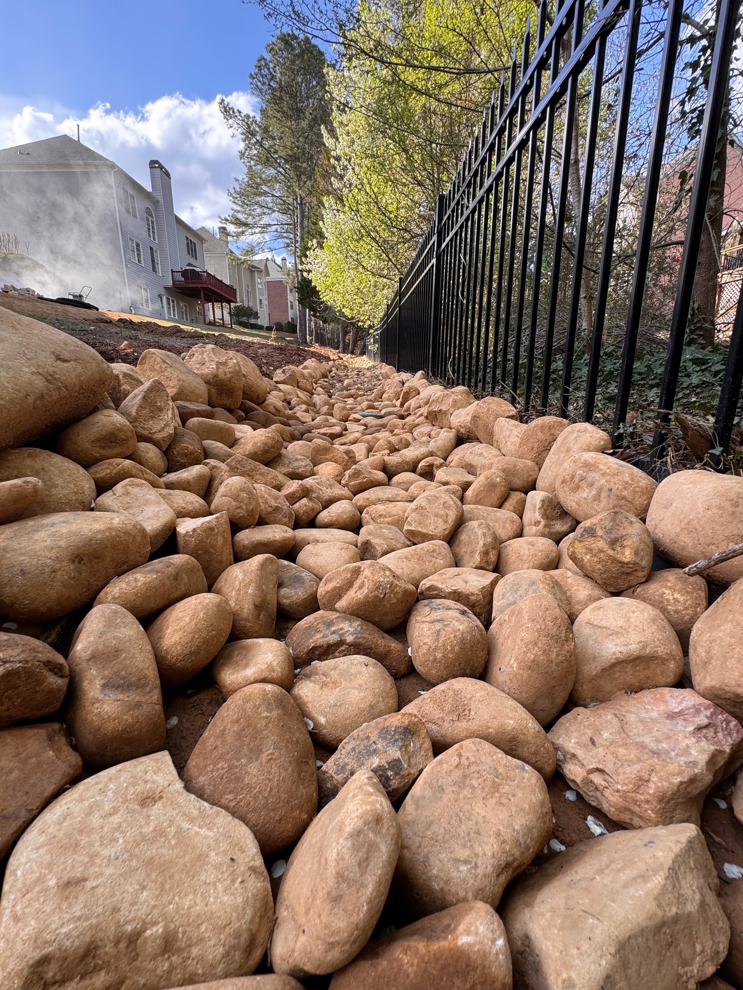 Smooth tan river rocks piled along a black metal fence in a landscaped backyard.