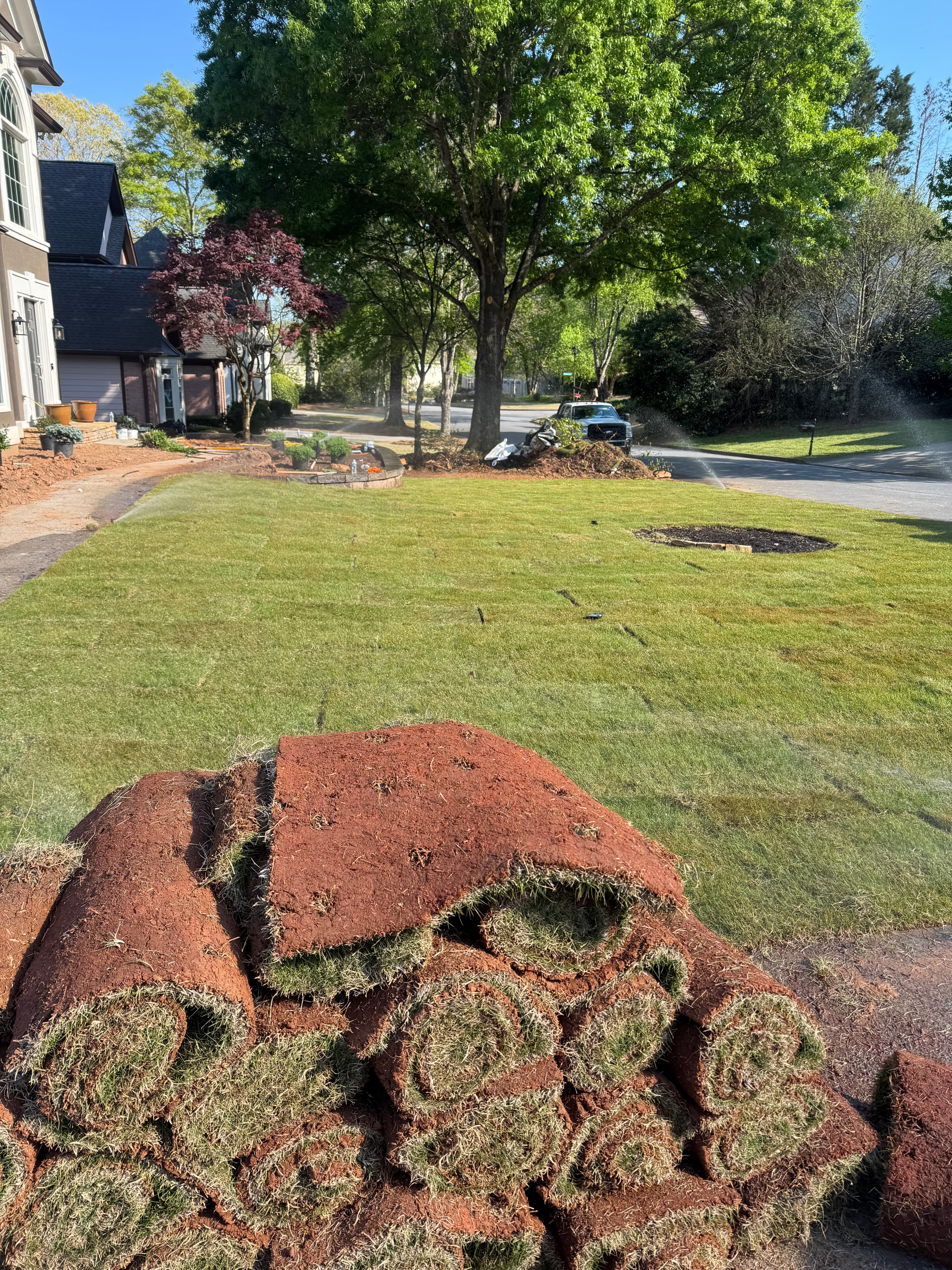 Stacked rolls of fresh sod beside a newly installed lawn in a sunny residential neighborhood.