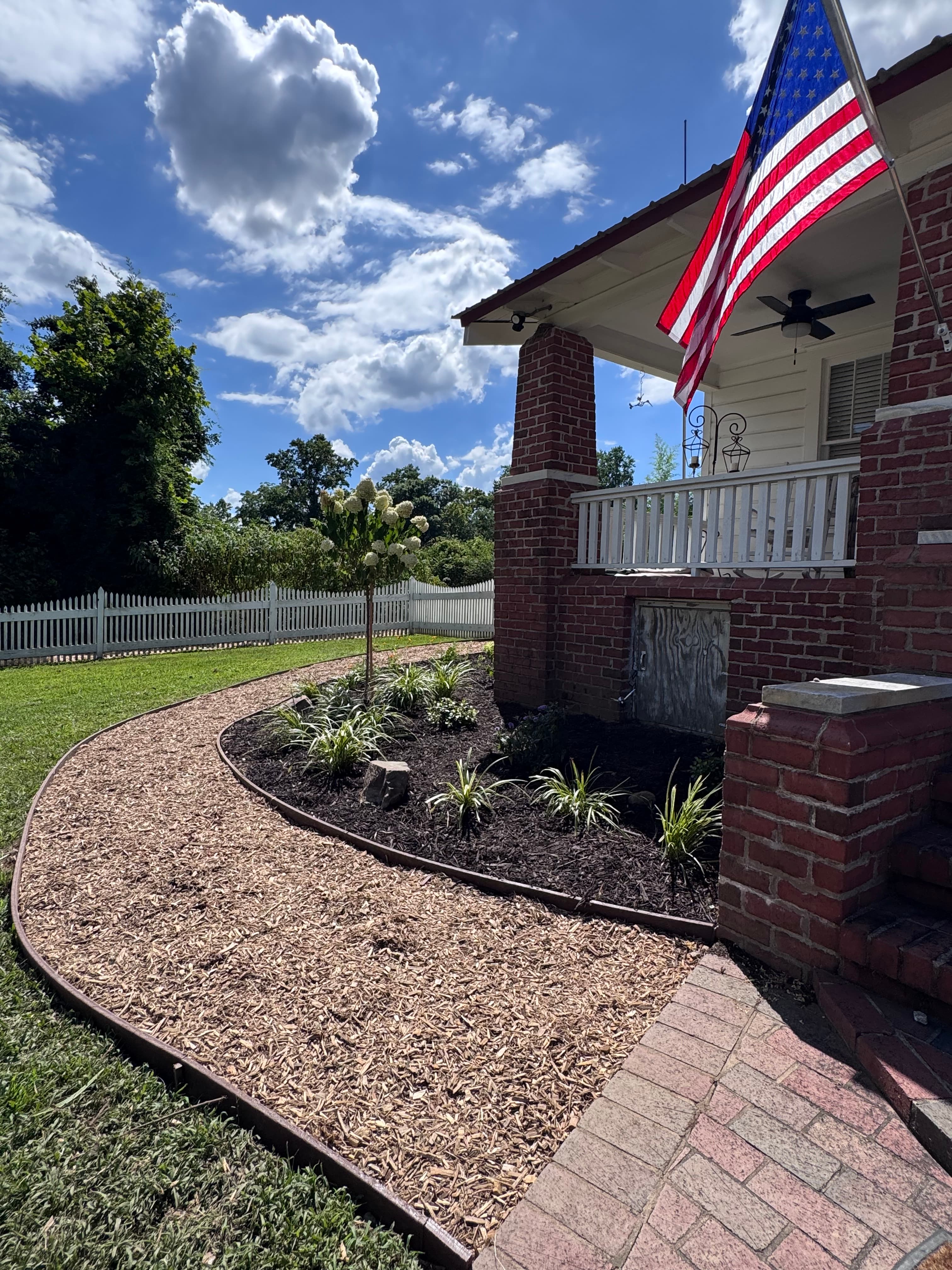 American flag on a brick porch overlooking a landscaped garden and white picket fence.