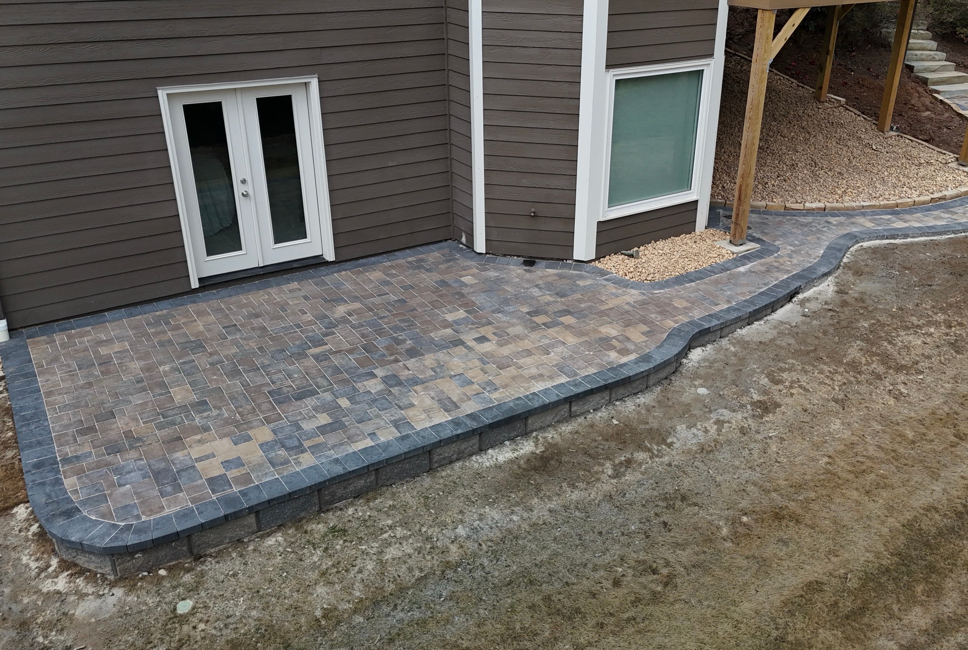 Stone paver patio and curved walkway with dark borders next to a brown house.