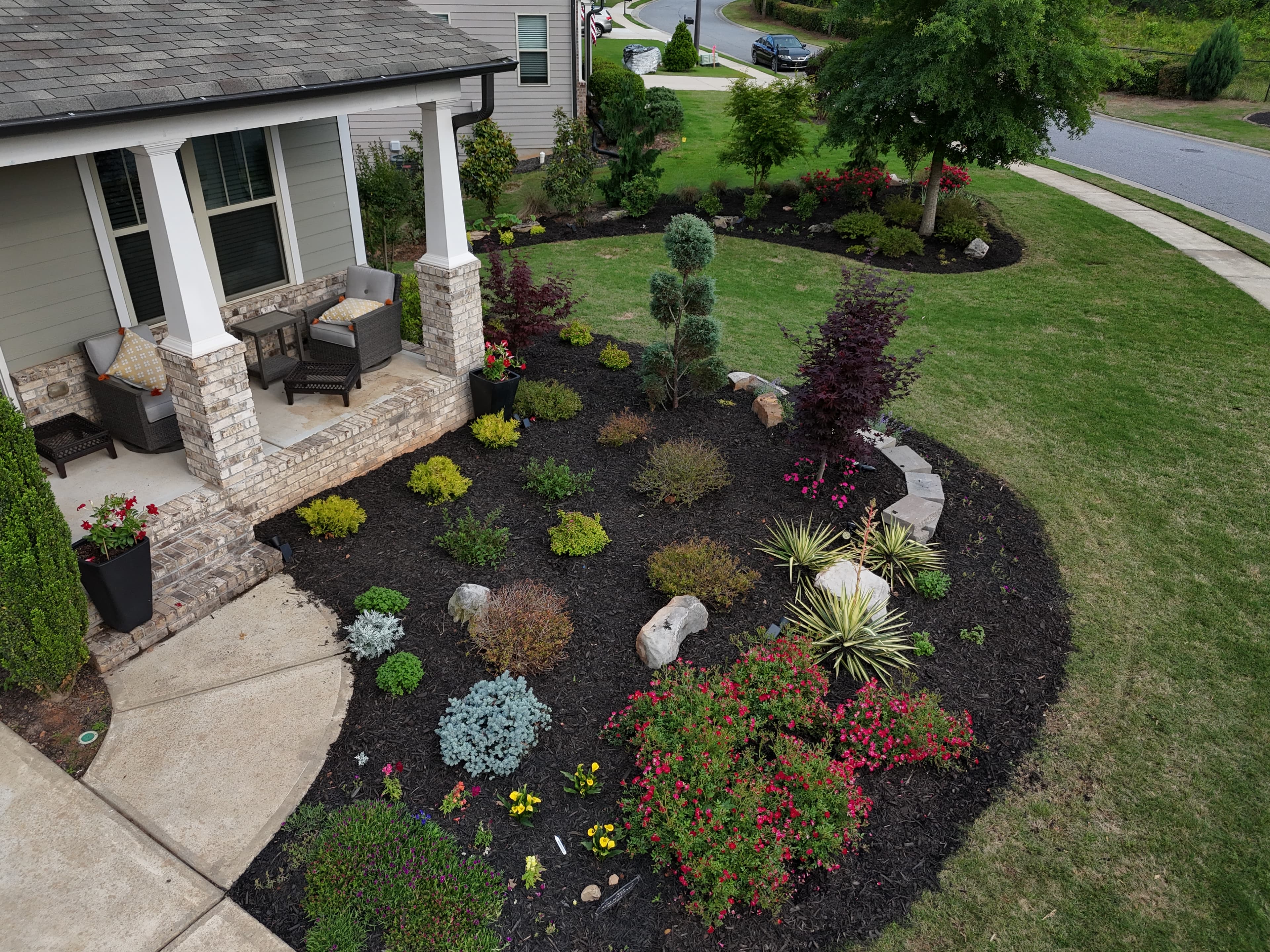 Front porch and a large, curved garden bed with colorful plants, shrubs, and dark mulch.